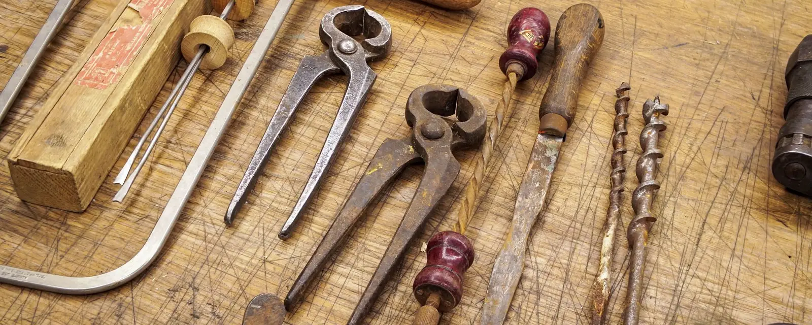 Pincers, chisels, drill bits and a hacksaw laid out on a wooden workbench